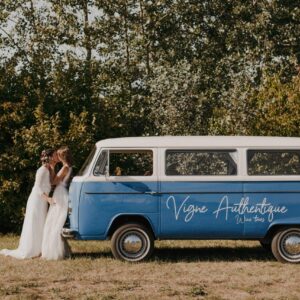 Deux mariées s'embrassent près d'un van vintage bleu et blanc Vigne Authentique Wine tours, dans un cadre naturel.