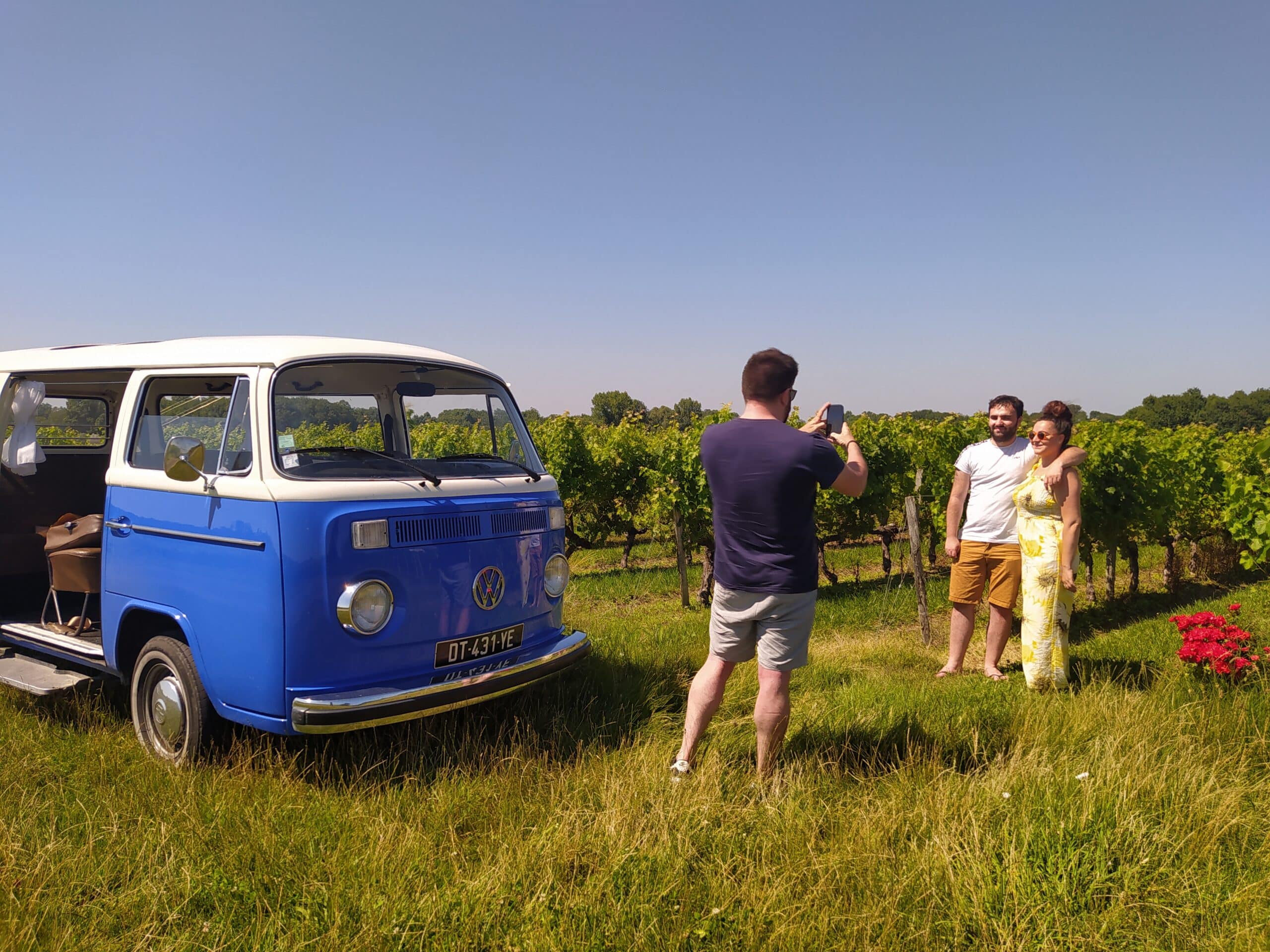 Combi Volkswagen bleu et blanc (DT-431-YE) dans un champ, un homme photographiant un couple souriant au milieu d'un vignoble.