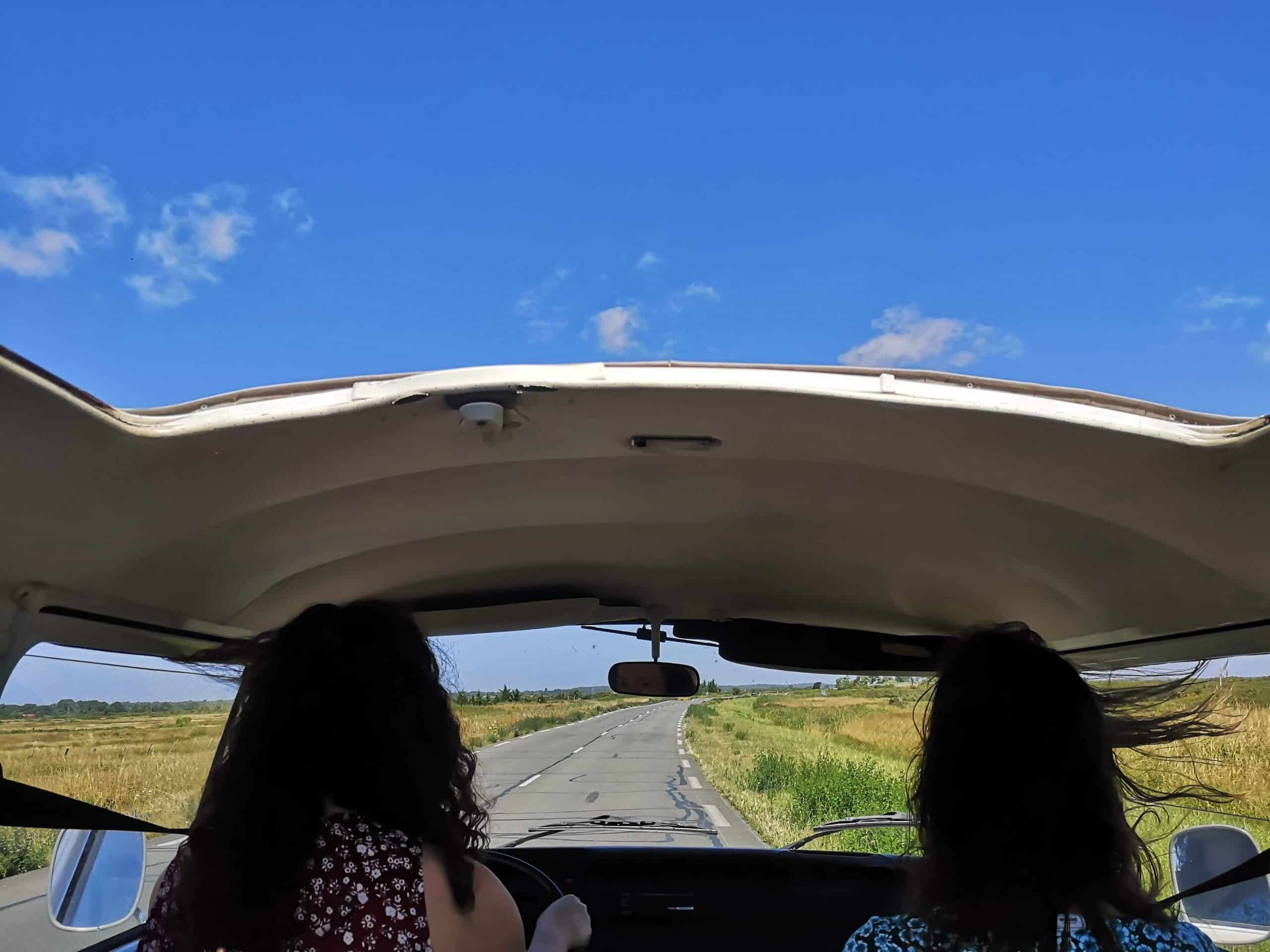 Deux femmes en van vintage, toit ouvert, sur route de campagne. Ciel bleu, champs, cheveux au vent. Une évasion libre en road trip.