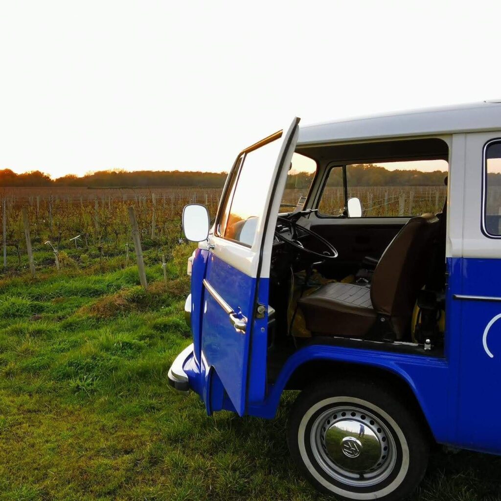 Van Combi VW: coucher de soleil magique dans un vignoble Van Combi VW bleu et blanc porte ouverte, au coucher du soleil dans un vignoble verdoyant. Ambiance de détente et road trip.
