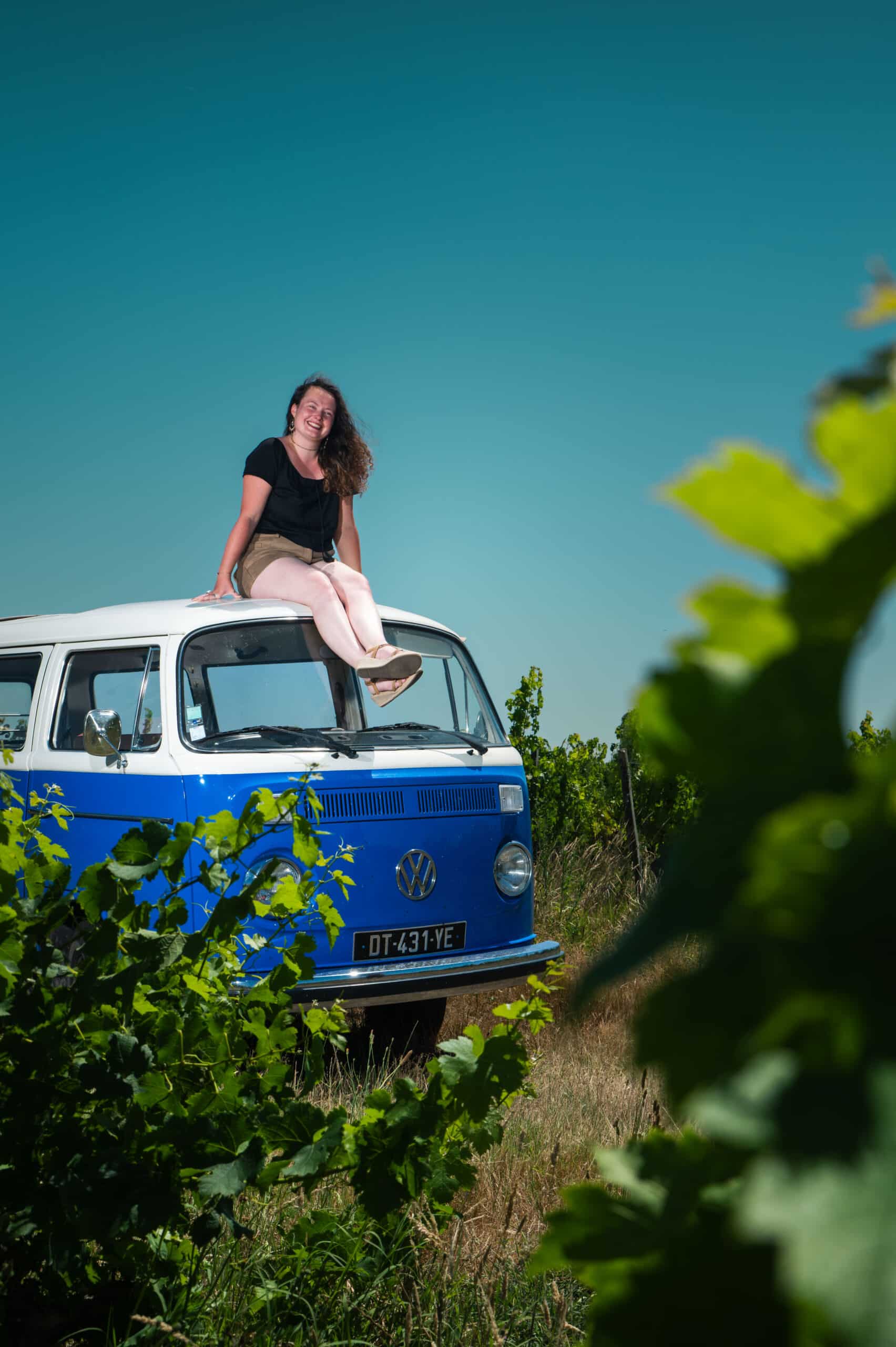 Jeune femme souriante assise sur le toit blanc et bleu d'un Combi Volkswagen immatriculé DT-431-YE, près d'une vigne.