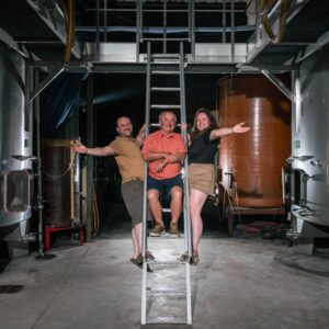 Trois personnes souriantes, deux hommes et une femme, posent sur une échelle au milieu d'une salle de cuves métalliques d'une cave.